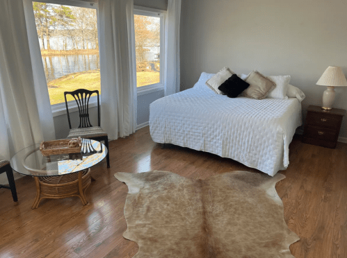 Cozy bedroom with a bed, a glass table, and a cowhide rug, featuring large windows overlooking a serene lake.