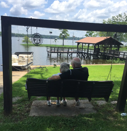 An elderly couple sits on a swing by a lake, enjoying the view and each other's company on a sunny day.