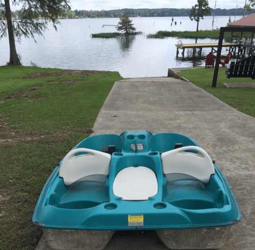 A turquoise paddle boat sits on a concrete ramp leading to a calm lake with trees and a dock in the background.
