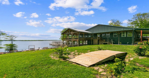 A lakeside view featuring a green building, wooden deck, and a grassy area under a blue sky with scattered clouds.