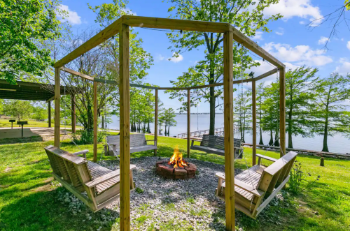 A cozy fire pit surrounded by wooden swings, set by a lake with trees and a clear blue sky in the background.