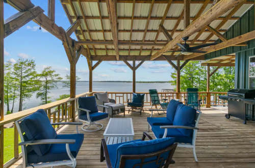 A spacious wooden deck with blue chairs and a table, overlooking a serene lake and green trees.