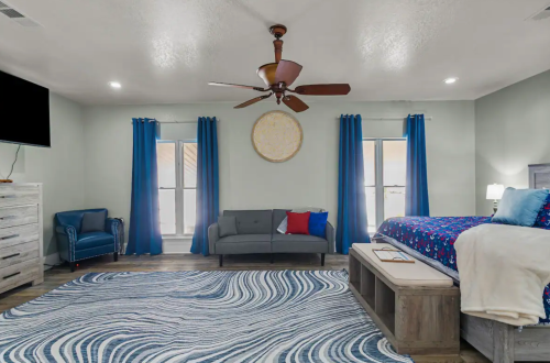 A spacious bedroom with a blue and white rug, a bed with colorful bedding, a sofa, and large windows with blue curtains.