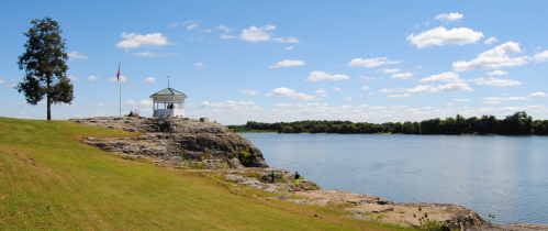 A scenic view of a river with a gazebo on a rocky shore, surrounded by green grass and blue skies with fluffy clouds.