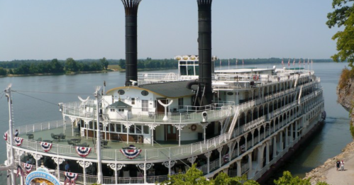 A large steamboat with two smokestacks docked by a river, decorated with flags and bunting.