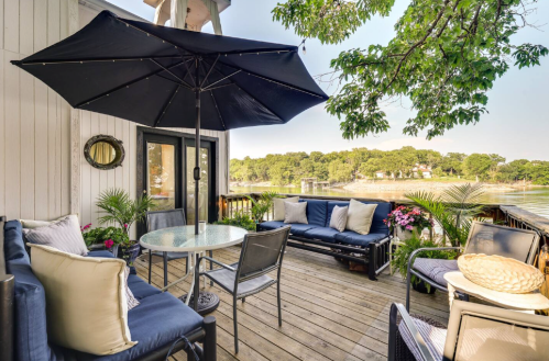 A cozy outdoor patio with a table, chairs, and an umbrella, overlooking a serene lake surrounded by trees.