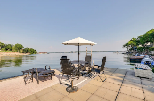 A lakeside patio with a table, chairs, and an umbrella, overlooking calm water and boats in the distance.