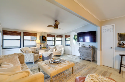 Cozy living room with light-colored furniture, a ceiling fan, and large windows, featuring a TV and decorative accents.