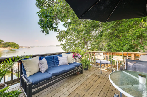 A cozy deck overlooking a serene lake, featuring a blue sofa, potted plants, and a table with chairs under a black umbrella.