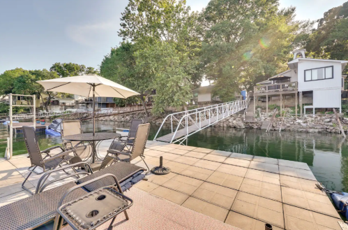 A lakeside patio with a table and chairs, an umbrella, and a bridge leading to a house across the water.
