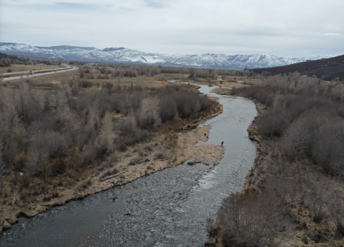 Aerial view of a winding river surrounded by bare trees and mountains, with a person fishing in the distance.