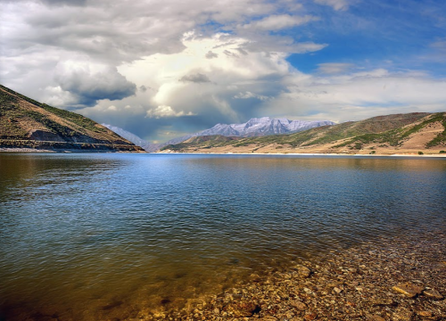 A serene lake surrounded by mountains under a cloudy sky, with clear water reflecting the landscape.