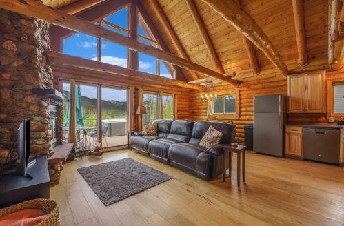 Cozy log cabin interior with a stone fireplace, large windows, and a comfortable sofa overlooking scenic views.