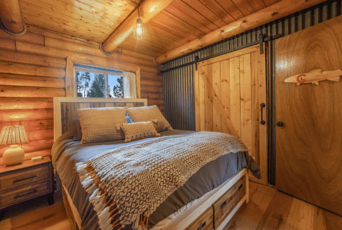 Cozy log cabin bedroom with a wooden bed, warm lighting, and rustic decor, featuring a window with forest views.