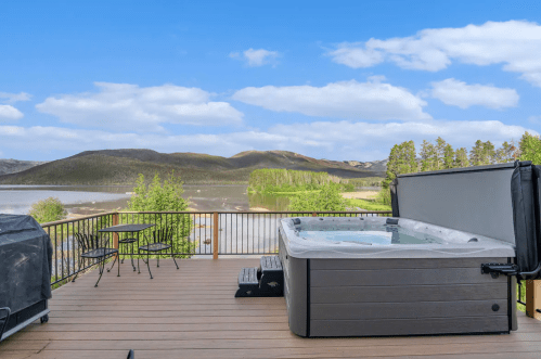 A scenic deck with a hot tub overlooking a lake and mountains under a blue sky with fluffy clouds.
