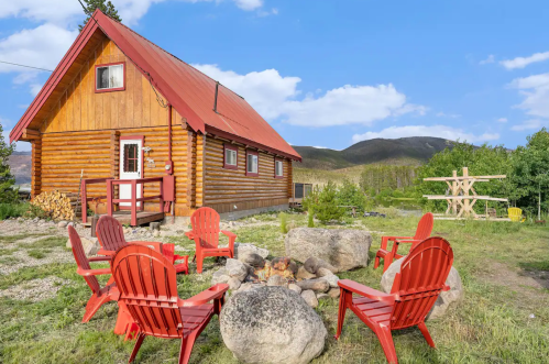 A cozy log cabin with a red roof, surrounded by a fire pit and Adirondack chairs, set against a scenic mountain backdrop.