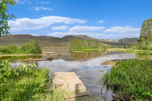A serene lake surrounded by greenery and mountains, with a wooden dock extending into the calm water under a blue sky.