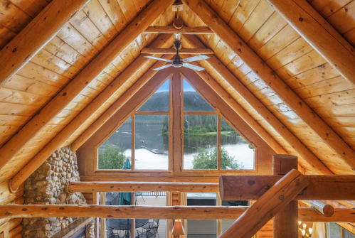 View from a wooden loft showcasing a triangular ceiling and large windows overlooking a serene lake and mountains.