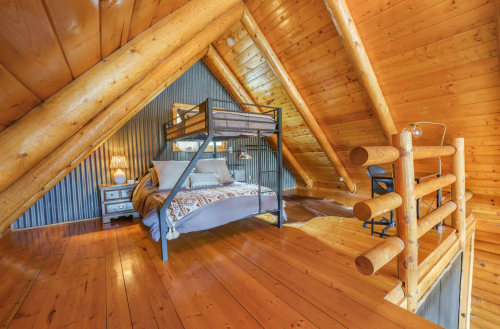 Cozy attic bedroom with a metal bunk bed, wooden beams, and warm lighting, featuring rustic decor and wooden flooring.