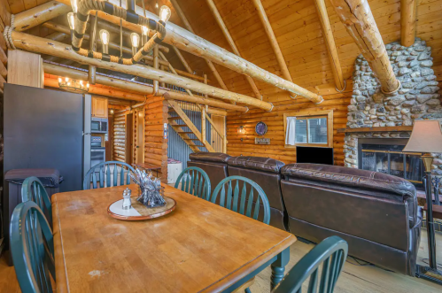 Cozy log cabin interior featuring a dining table, leather couches, and a stone fireplace with wooden beams overhead.
