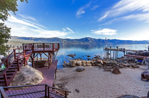 Scenic lakeside view with wooden decks, rocky shore, and mountains under a clear blue sky.
