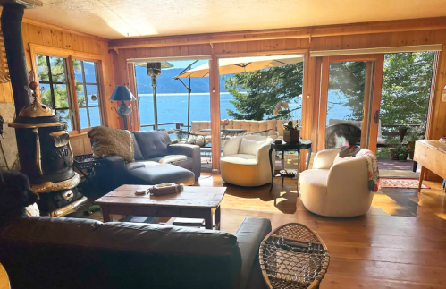 Cozy living room with wooden walls, comfortable seating, and large windows overlooking a lake and outdoor patio.