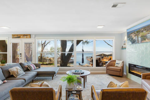 Cozy living room with a large sofa, coffee table, and a view of a lake through large windows. Modern decor and fireplace.