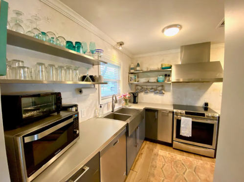 Modern kitchen with stainless steel appliances, open shelves, glassware, and a large sink. Bright and well-organized space.