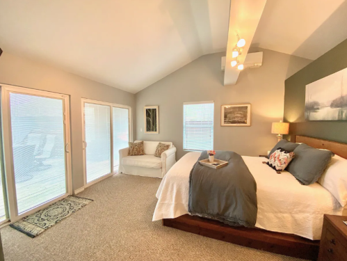 Cozy bedroom with a large bed, a white chair, and sliding glass doors leading to a deck. Soft lighting and neutral decor.