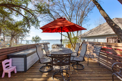 A lakeside deck with a round table, chairs, and a red umbrella, surrounded by trees and a view of the water.
