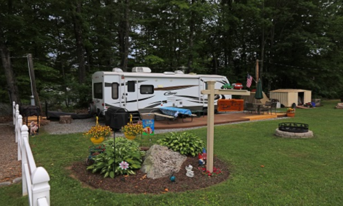A cozy RV parked in a green yard with a fire pit, garden decorations, and a picnic table under trees.