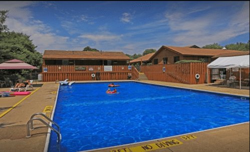 A sunny pool area with people relaxing, surrounded by wooden buildings and trees under a blue sky.