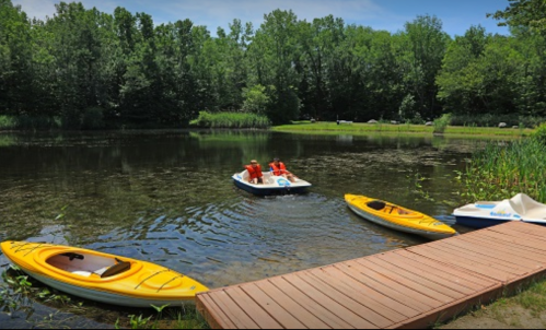 A serene pond surrounded by trees, with yellow kayaks and a small boat on the water, and a wooden dock nearby.