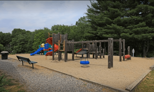 A playground with slides, swings, and climbing structures, surrounded by trees and gravel paths.