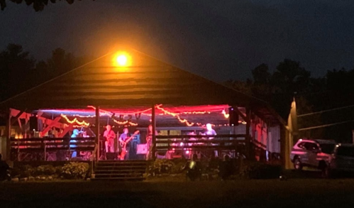 A band performs on a brightly lit outdoor stage at night, with colorful lights and a rustic venue.
