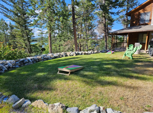 A grassy yard with stone borders, two cornhole boards, and green chairs, surrounded by trees and a lake in the background.