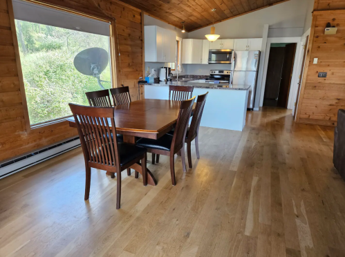 A wooden dining table with chairs in a spacious room, featuring a kitchen area and large windows with greenery outside.