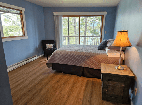 Cozy bedroom with a large window, a bed, a chair, and a wooden nightstand, featuring blue walls and natural light.
