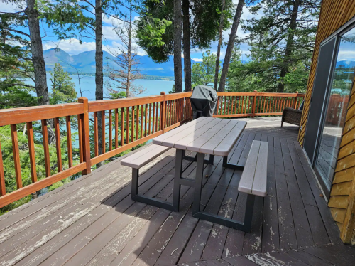 A wooden deck with a picnic table and grill, surrounded by trees and overlooking a lake and mountains in the distance.