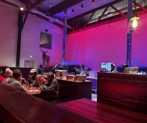 A cozy restaurant interior with dim lighting, featuring diners at wooden tables and a visible kitchen area in the background.