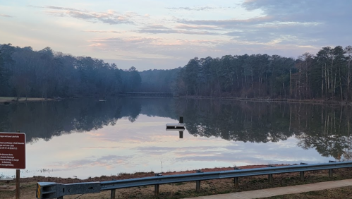 A calm lake reflecting trees and a cloudy sky, with a small dock visible in the water.