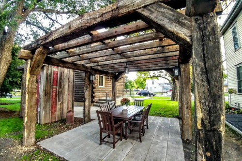 A rustic outdoor patio with a wooden pergola, featuring a dining table and chairs surrounded by greenery.