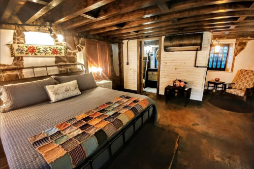 Cozy bedroom with a quilted bed, wooden beams, and rustic decor, featuring a small sitting area and a bathroom door.