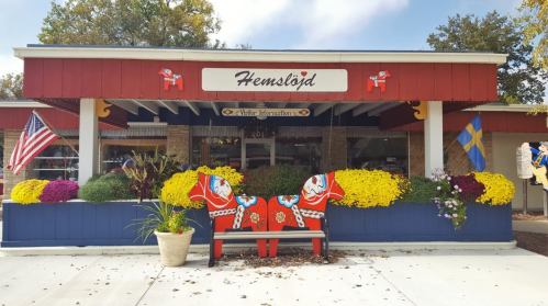 A colorful storefront with a sign reading "Hemslöjd," featuring flower displays and decorative horse sculptures.
