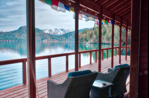 A serene lakeside view from a wooden deck, featuring chairs and colorful prayer flags against a backdrop of mountains.