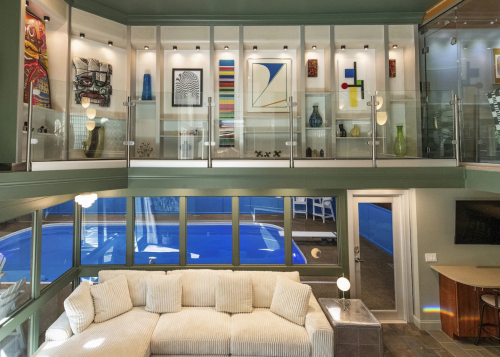 Modern living room with a large sectional sofa, glass walls, and artwork, overlooking a pool area.