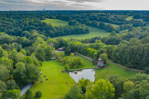 Aerial view of lush green fields, a pond, and a small red building surrounded by trees under a cloudy sky.