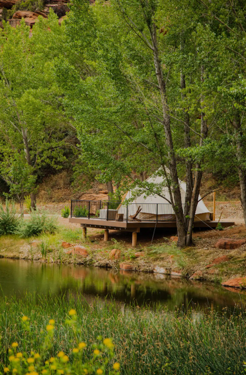 A cozy tent on a wooden deck by a serene pond, surrounded by lush green trees and natural scenery.