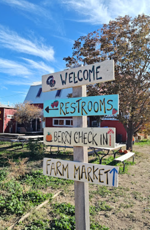 A wooden signpost with directional arrows for restrooms, berry check-in, farm market, and a welcome message.