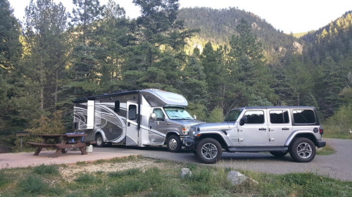 An RV parked beside a Jeep at a campsite, surrounded by trees and mountains in the background.